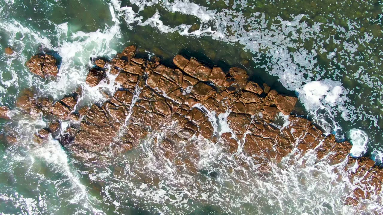 Top-down La Bocana shoreline view with whitewater curling around scattered rocks and a larger brown formation, with no visible people.