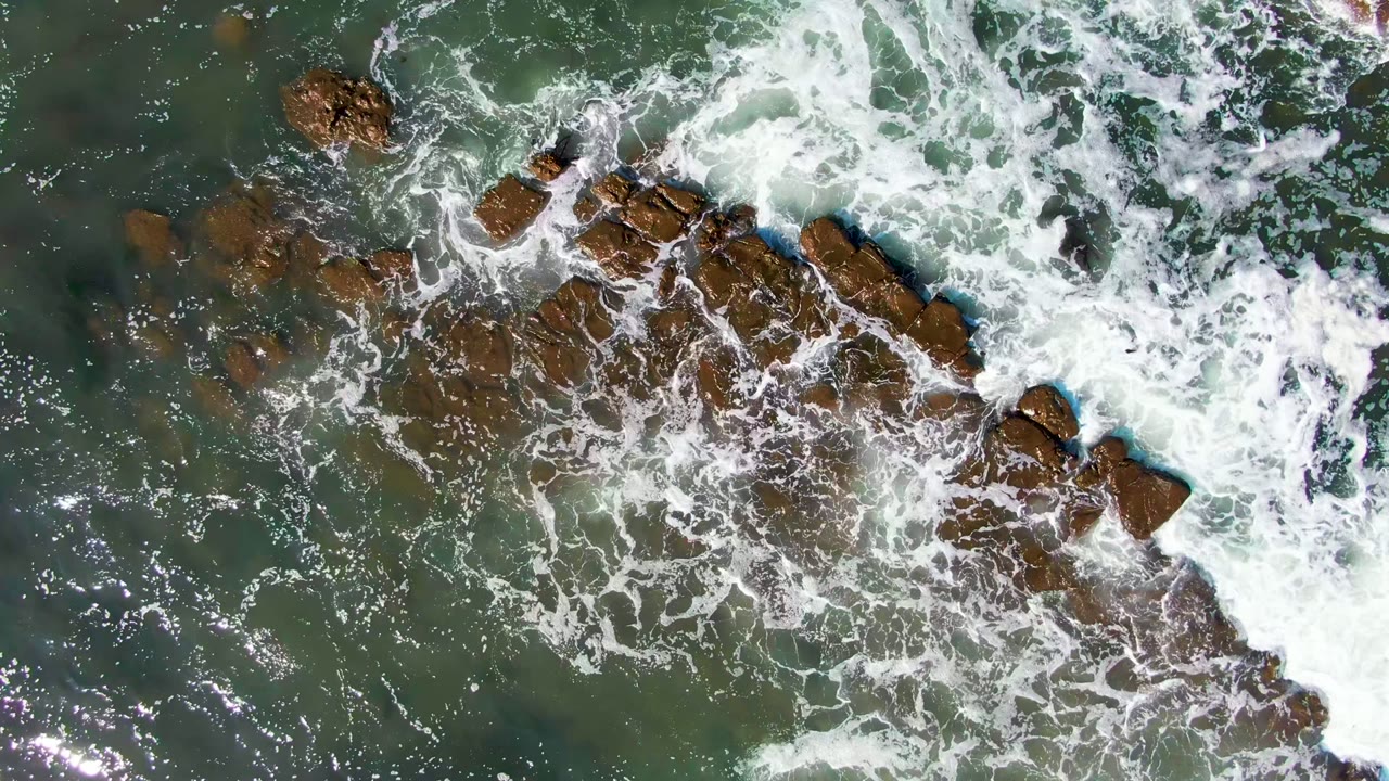 Overhead view in La Bocana, Baja California Sur, with white surf spreading over brown rock outcrops in green water and no visible people.