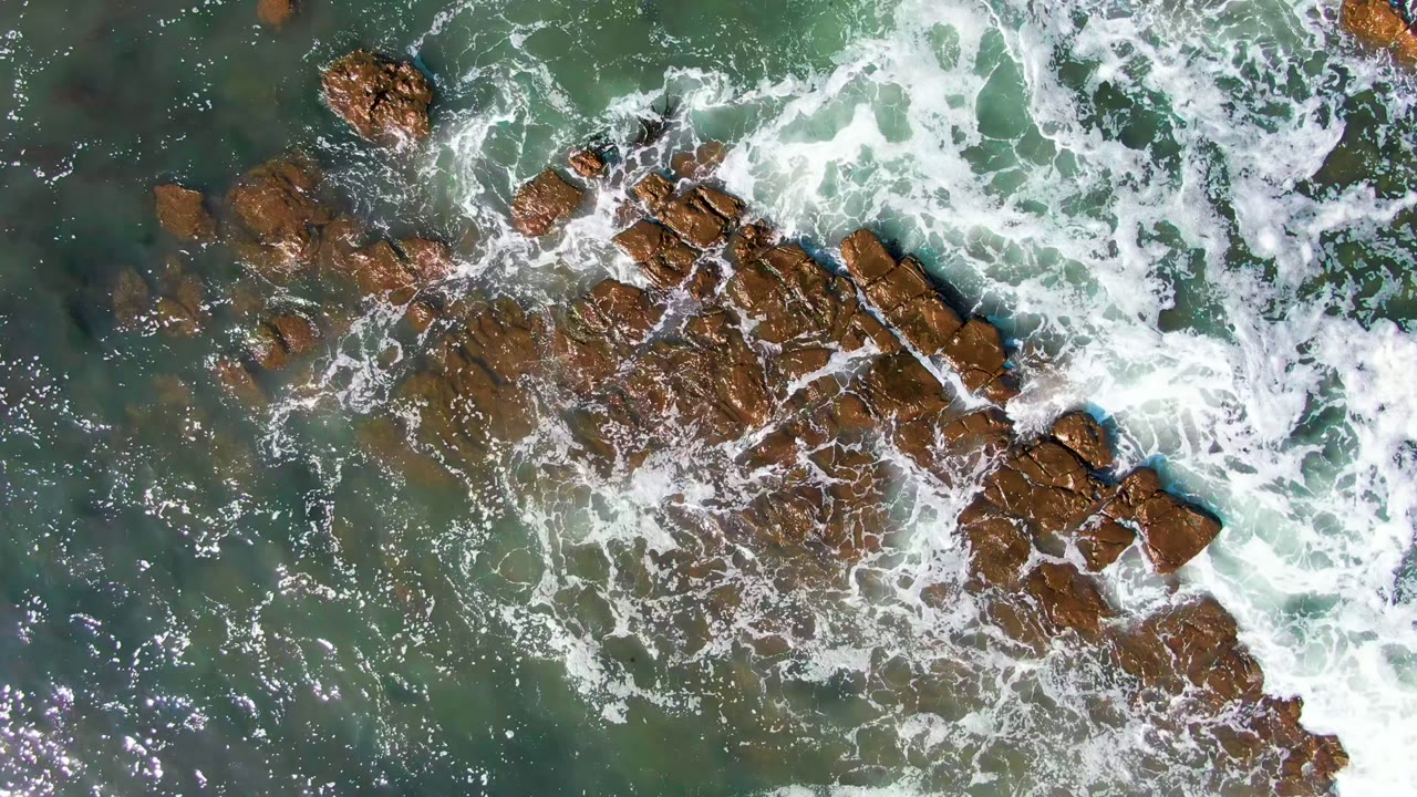 Top-down view in La Bocana, Baja California Sur, with white waves breaking across diagonal rust-brown rocks and darker green water around them, with no visible people.