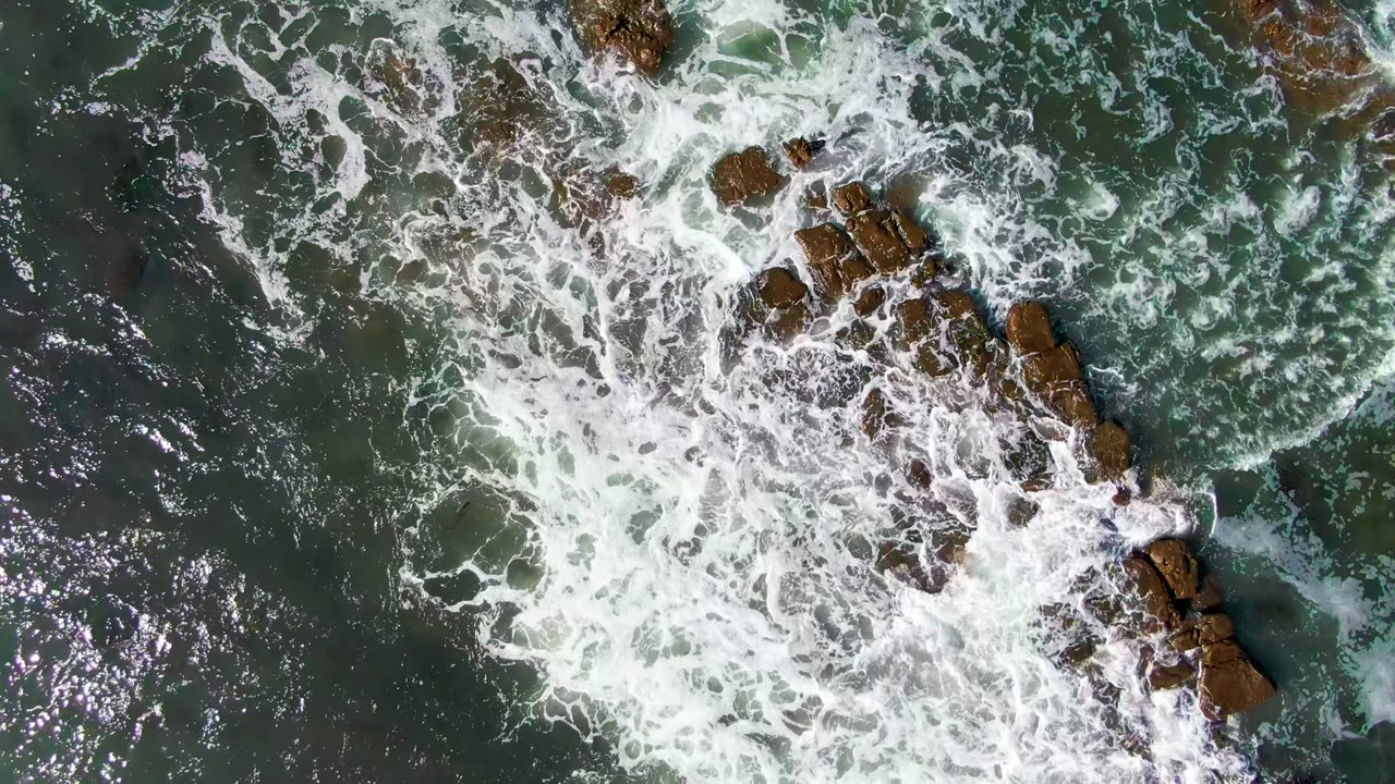 Overhead view in La Bocana, Baja California Sur, where white surf spreads over brown rock shelves in green ocean water, with no visible people.