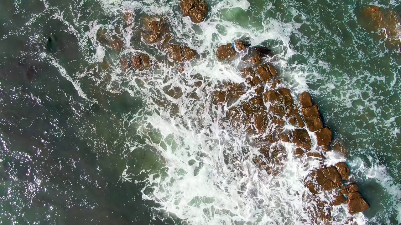 Top-down La Bocana coastal view with exposed brown rocks near the upper right and white foam breaking across green water, with no visible people.