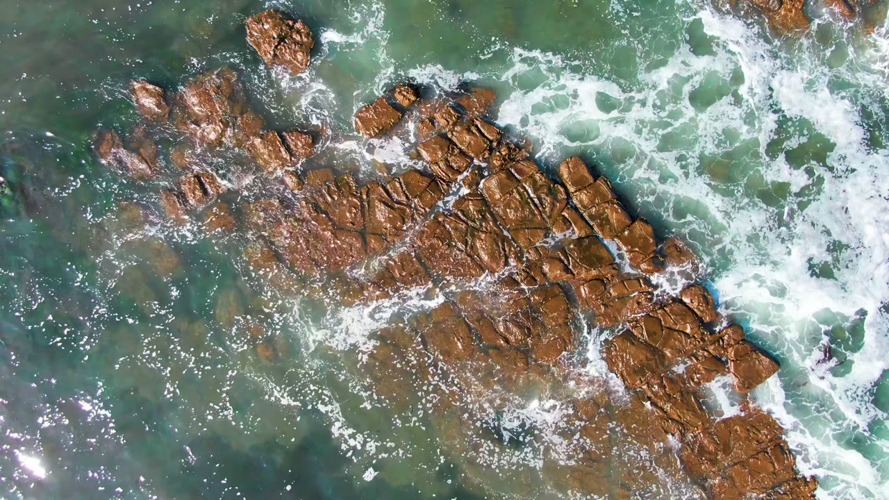 Top-down view in La Bocana, Baja California Sur of brown rock formations surrounded by turquoise-green water and scattered white foam, with no visible people.