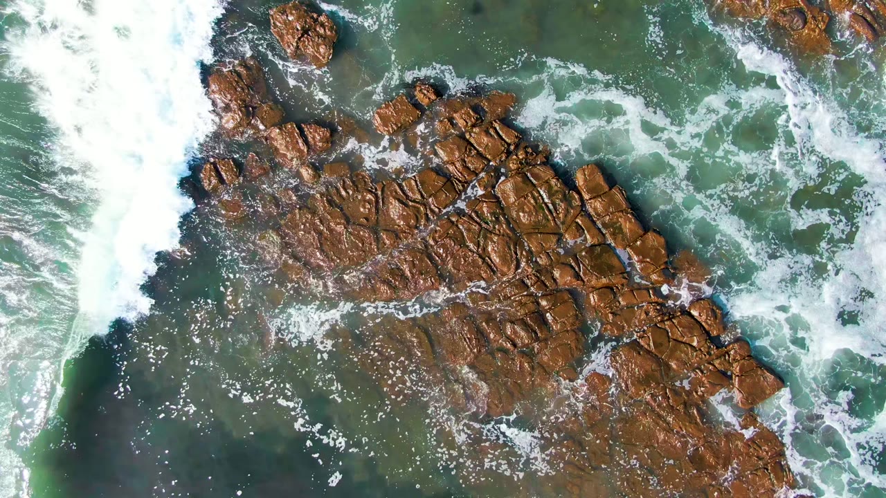 Overhead La Bocana coastline view with exposed brown rocks at center and a thick band of whitewater breaking along the right side, with no visible people.