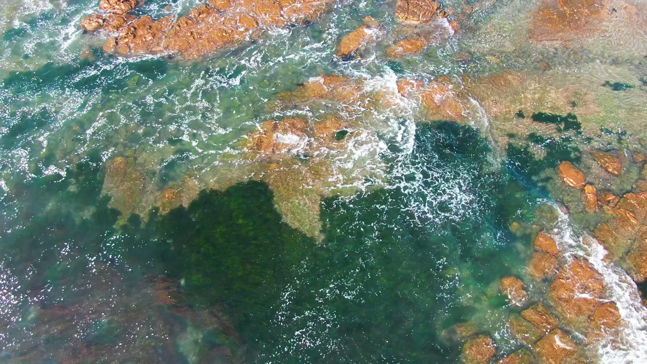 Overhead frame from La Bocana, Baja California Sur showing clear green-blue water washing over orange-brown coastal rocks with scattered white foam and no visible people.