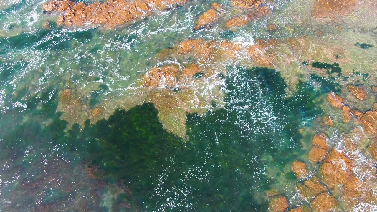 Aerial rocky shallows with green water and small foam streaks.