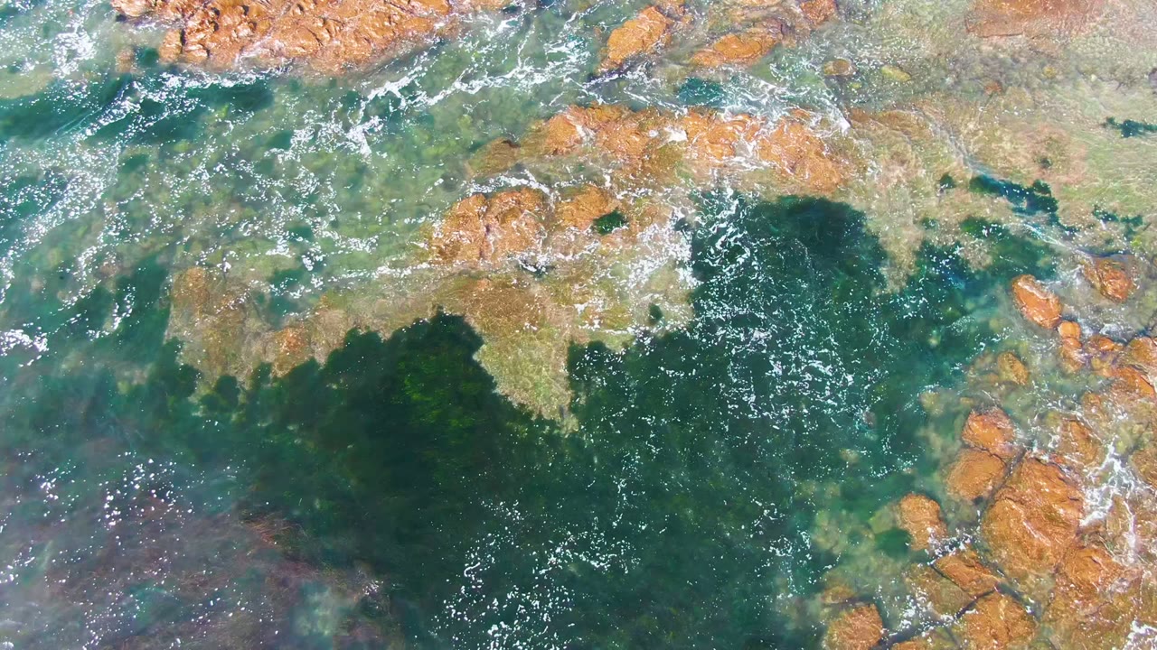 Overhead footage in La Bocana shows white foam curling around exposed orange rocks at the upper edge while darker green water fills the lower portion, with no visible people.
