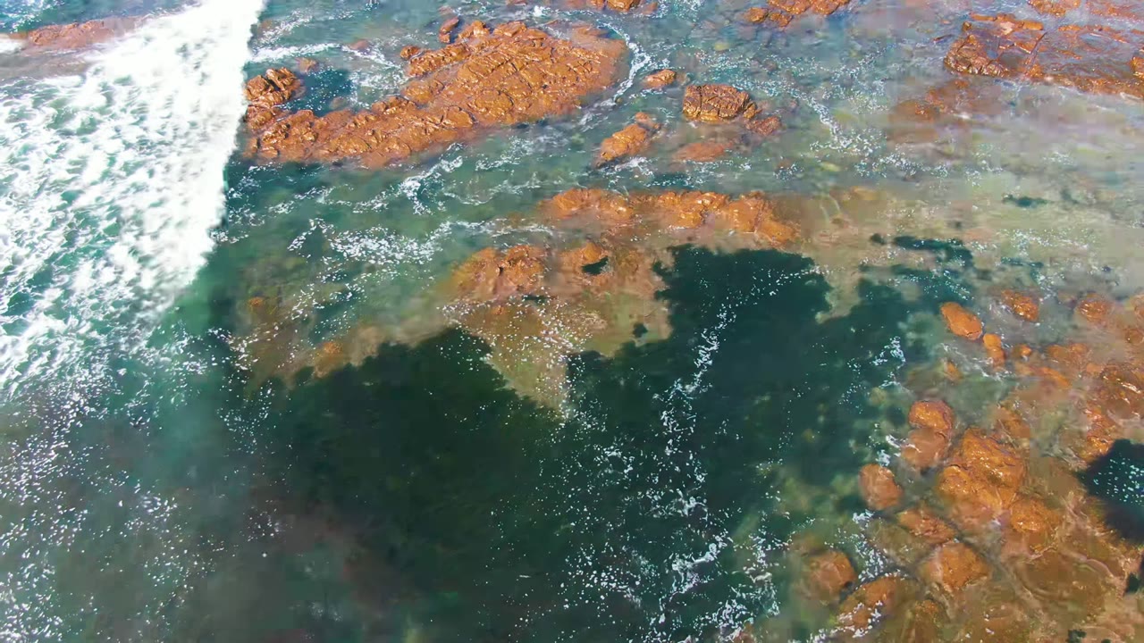 Overhead view in La Bocana, Baja California Sur showing white surf entering from the upper left over brown rocks and clear green water, with no visible people.