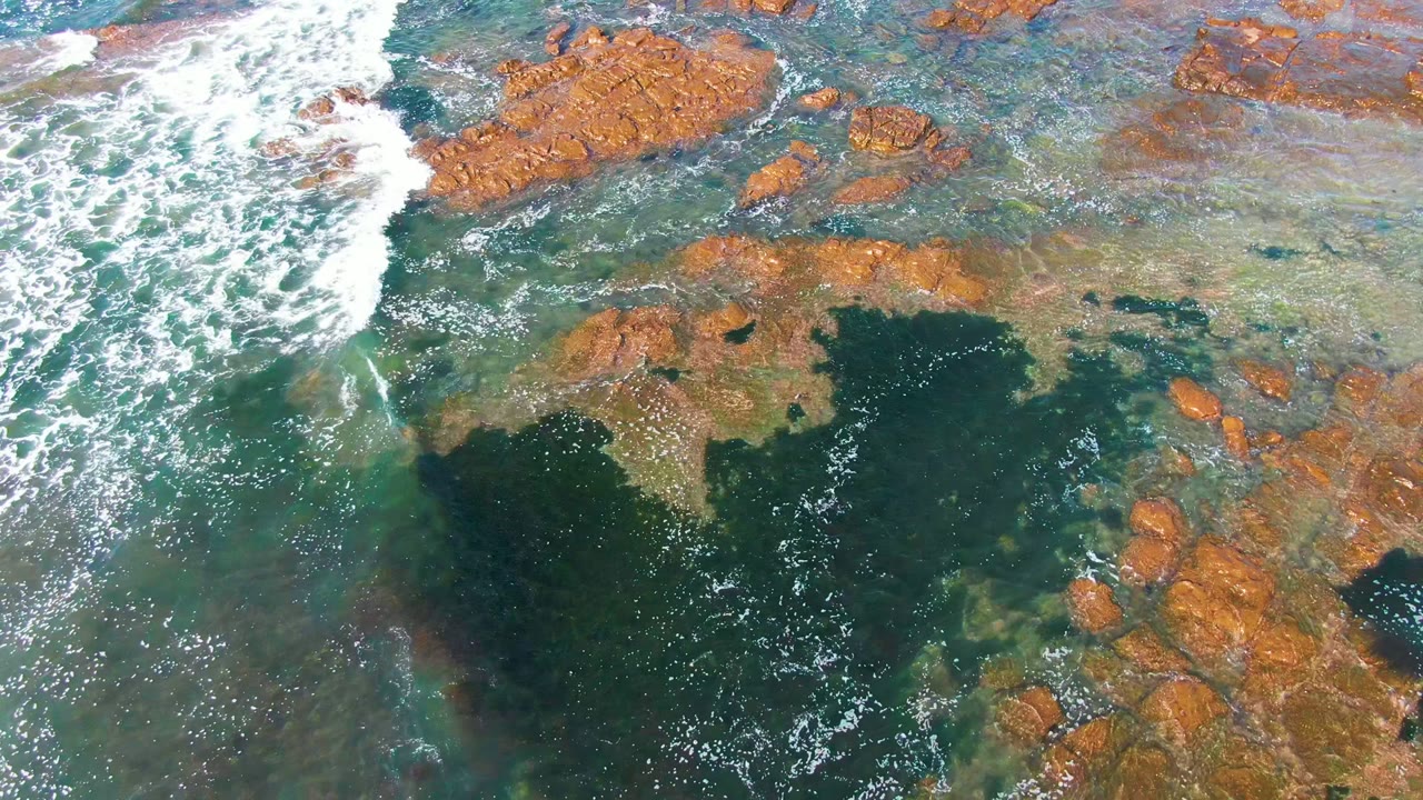 White wave foam spreads over shallow turquoise water and rocky reef.