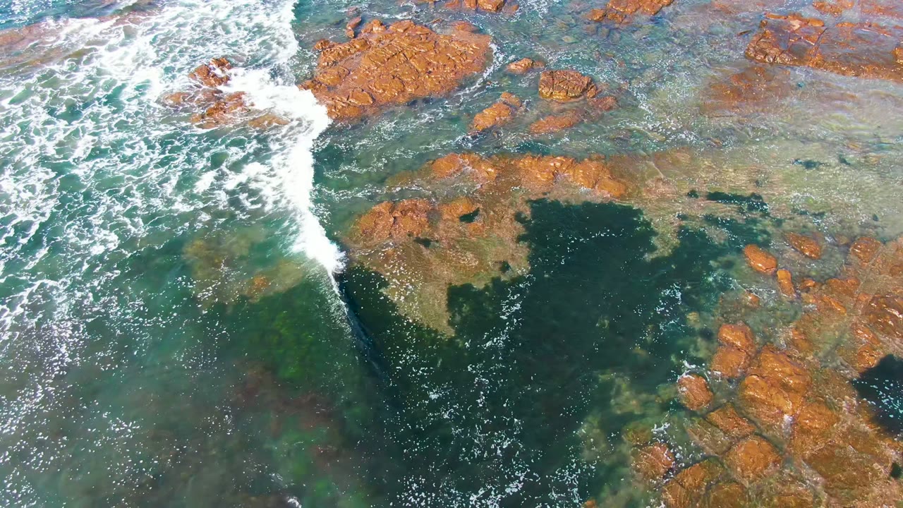 Top-down La Bocana coastline view where a diagonal strip of surf rolls over transparent water toward clustered brown rocks, with no visible people.