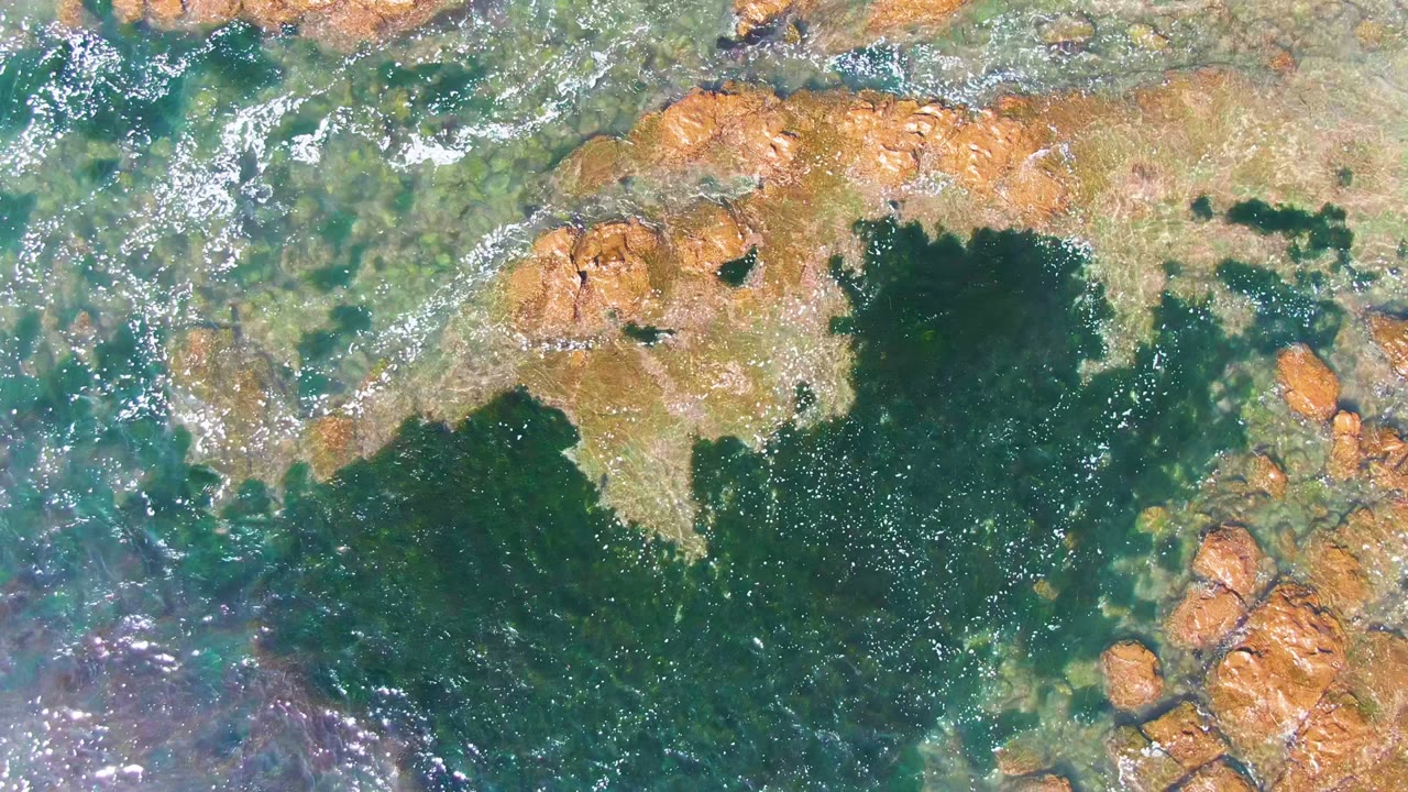 Foamy water crosses shallow rocks in a top-down coastal view.