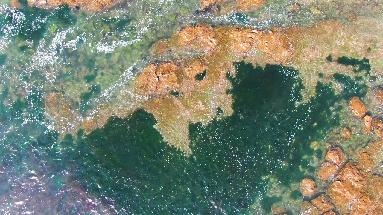 Aerial La Bocana frame where transparent blue-green water exposes rocks around a darker green pool, with no visible people.