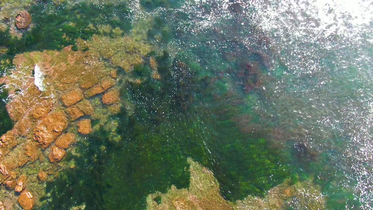 Overhead La Bocana frame showing green-blue water, muted brown rocks below the surface, a bright band of sun reflection across the upper right, and no visible people.