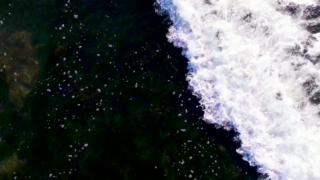 Overhead view in La Bocana, Baja California Sur, showing a thick white wave-foam band crossing dark green ocean water with no visible people.