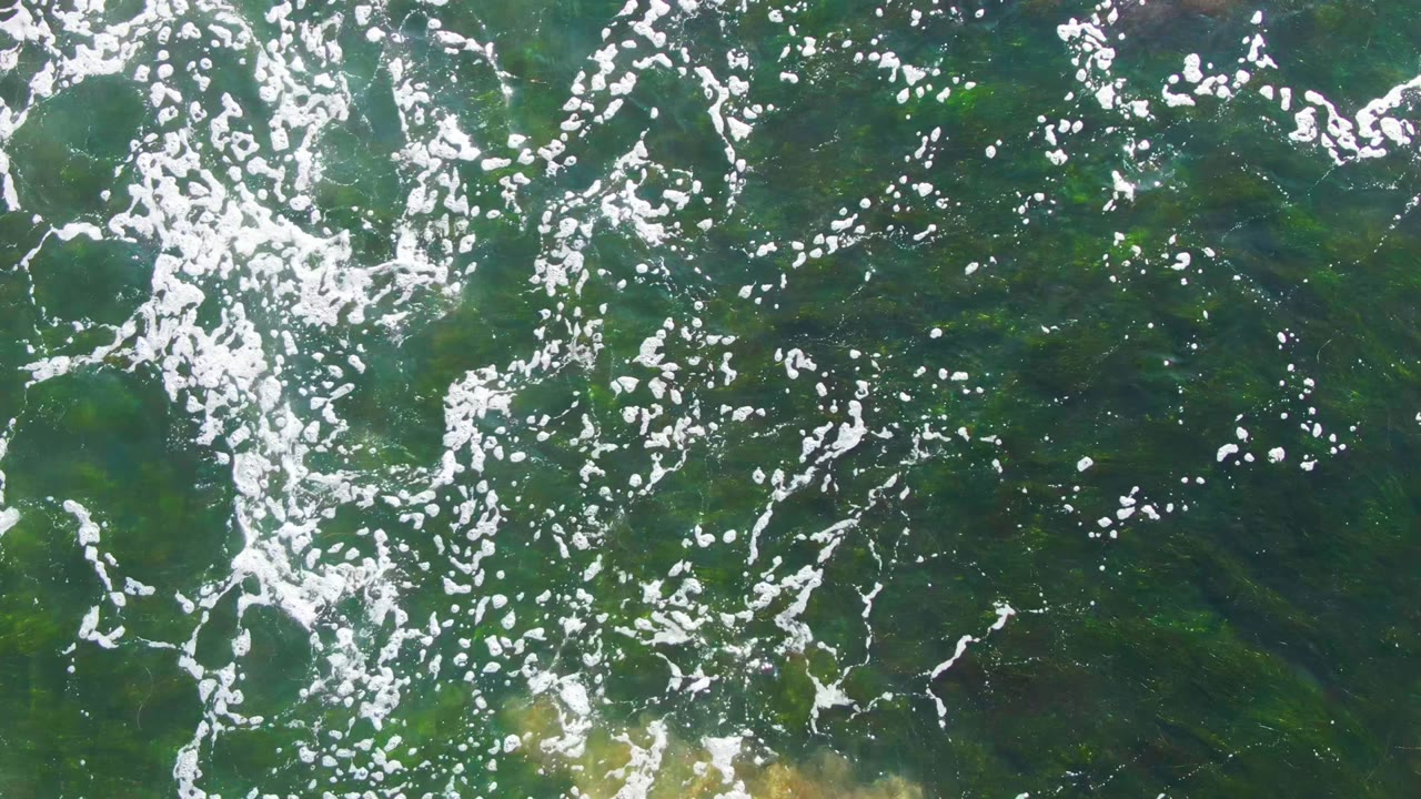Overhead frame of shallow green-blue water at La Bocana with scattered white sunlight glints, faint sandy seabed patches below the surface, and no visible people.