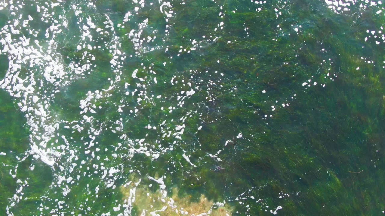 Clear shallow water at La Bocana fills the frame with dense white reflections near the left side, green submerged texture, sandy seabed edges, and no visible people.