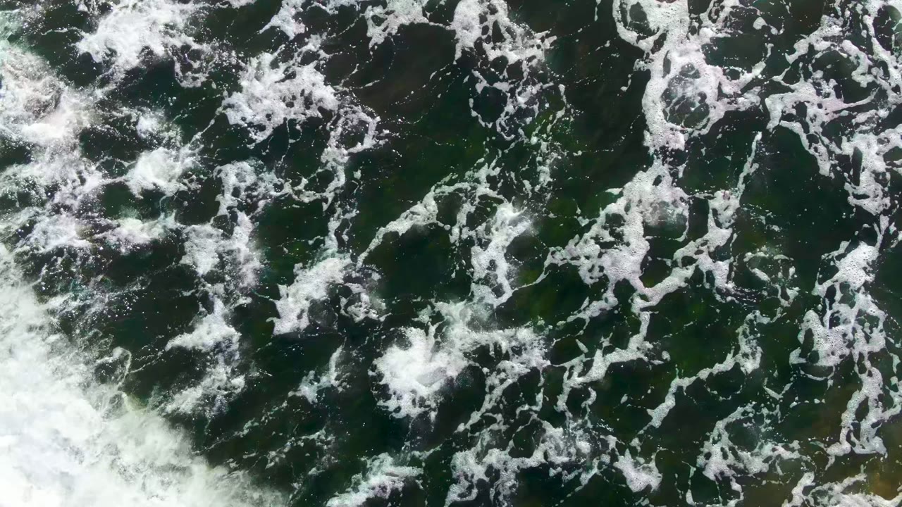 Overhead view of white wave foam spread across dark green ocean water in La Bocana, Baja California Sur, with no visible people.