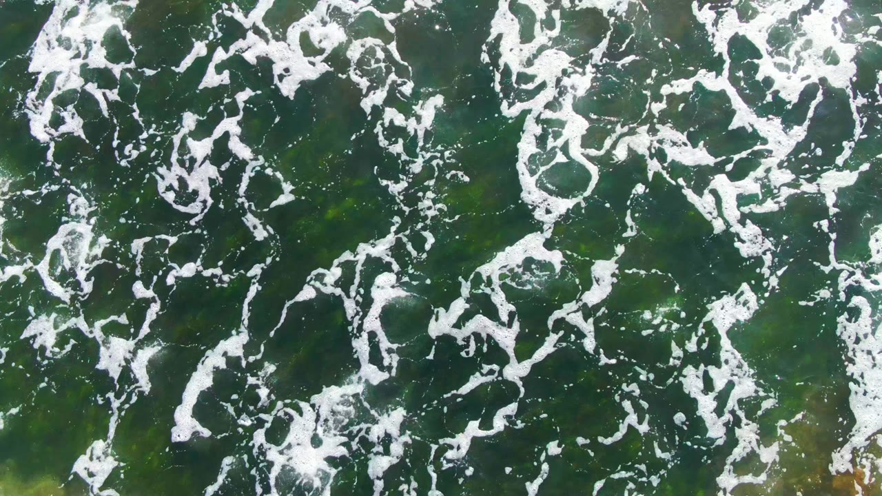 White foam streaks drift across green ocean water in La Bocana, Baja California Sur, with faint yellow-green tones below the surface and no visible people.