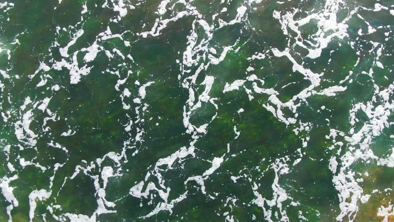 Scattered white foam lines sit over green water in La Bocana, Baja California Sur, with darker patches beneath the surface and no visible people.