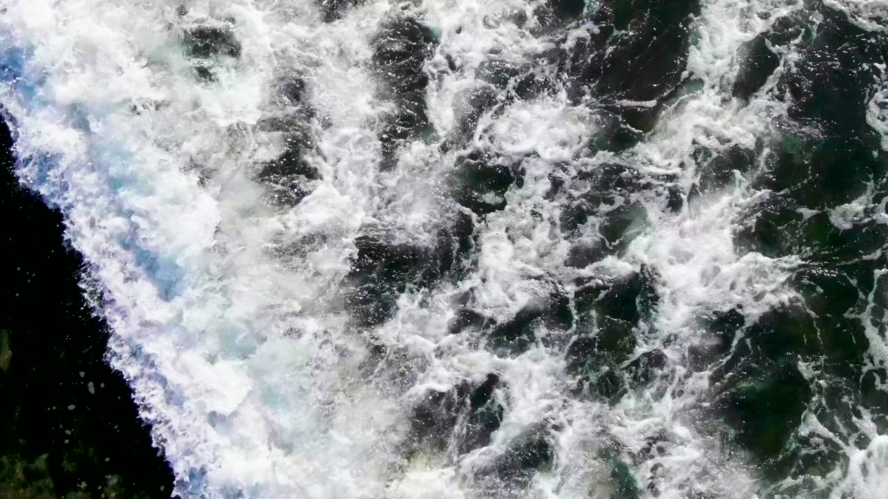 Overhead La Bocana frame showing churning white foam patterns across green coastal water, with darker pockets of water visible between the bubbles and no visible people.