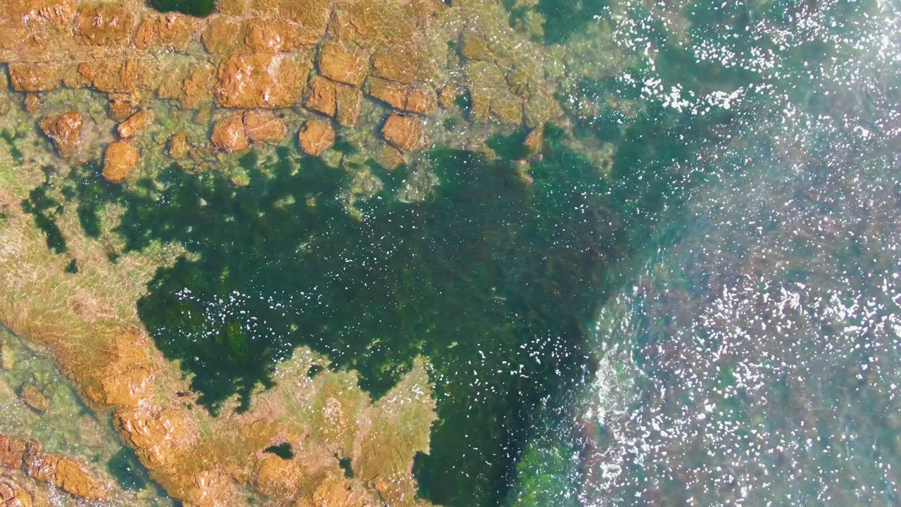 Overhead view of La Bocana shoreline with blocky brown rocks at the top, a dark tidal pool below them, clear turquoise-green water to the right, sun sparkle, and no visible people.