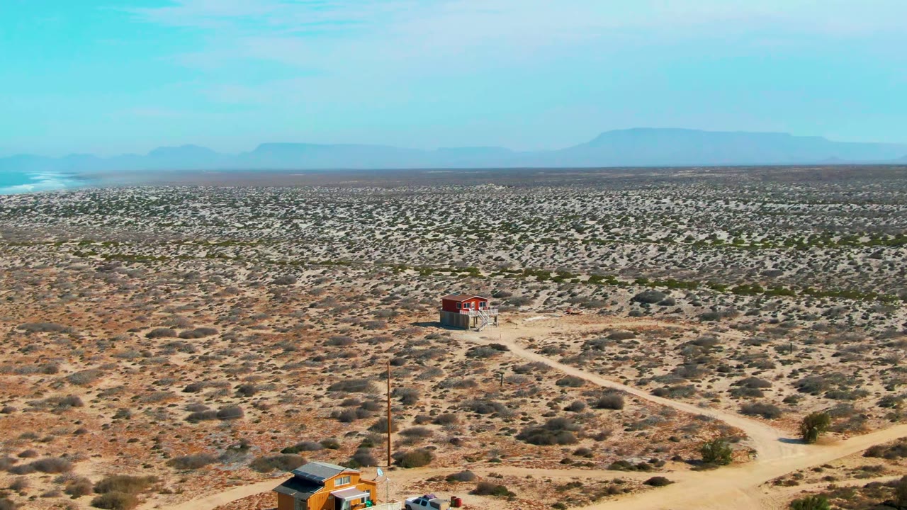 Wide aerial view in La Bocana, Baja California Sur, showing scrubby desert, a small red-roofed structure near dirt tracks, distant mountains, and partial foreground activity at the lower edge.
