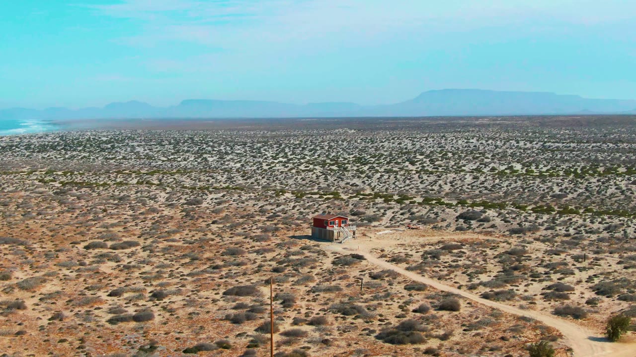 La Bocana desert scrub fills the frame as a small red-roofed structure sits near a branching dirt road, with a flat horizon and mountains under a clear blue sky.