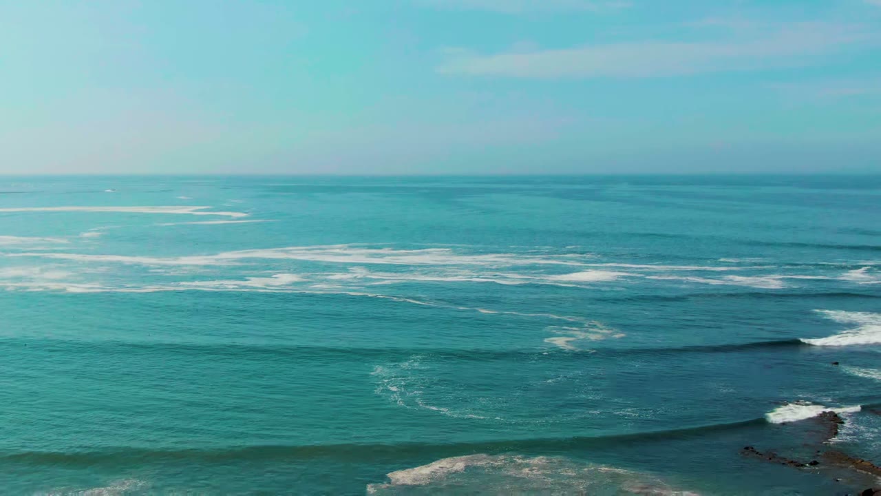 Wide aerial view of La Bocana ocean water with white surf bands crossing the frame, rocky shoreline just entering at the lower right, and no visible people.
