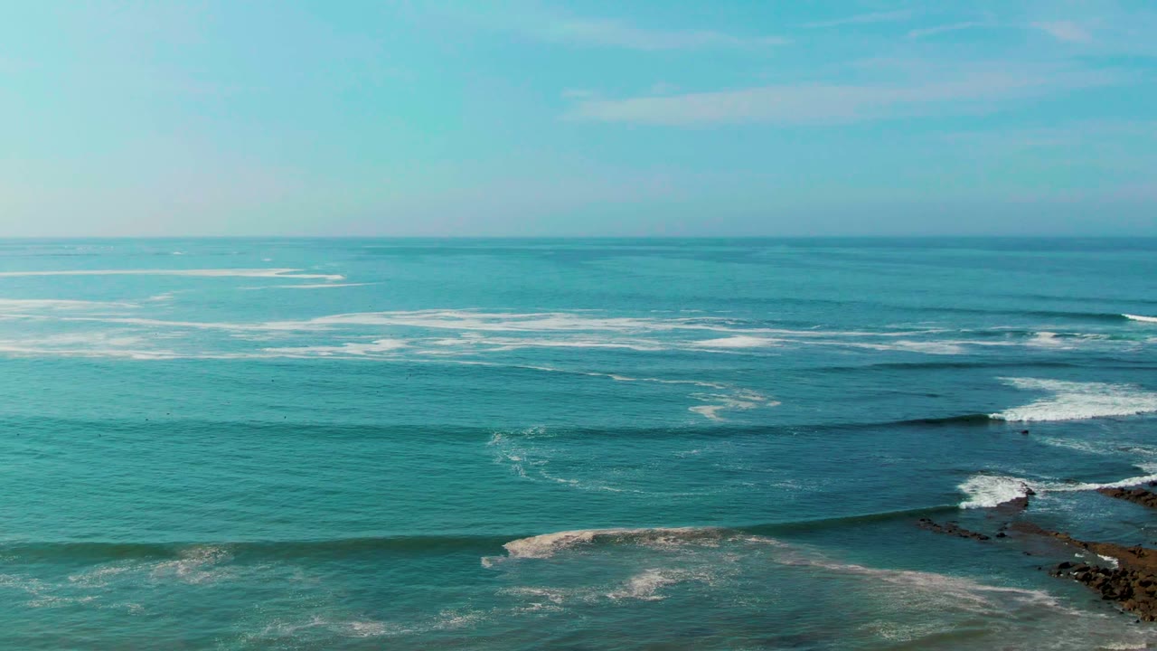 A long dark wave line moves across the La Bocana water, with white foam patches beyond it, rocks at the right edge, and no visible people.
