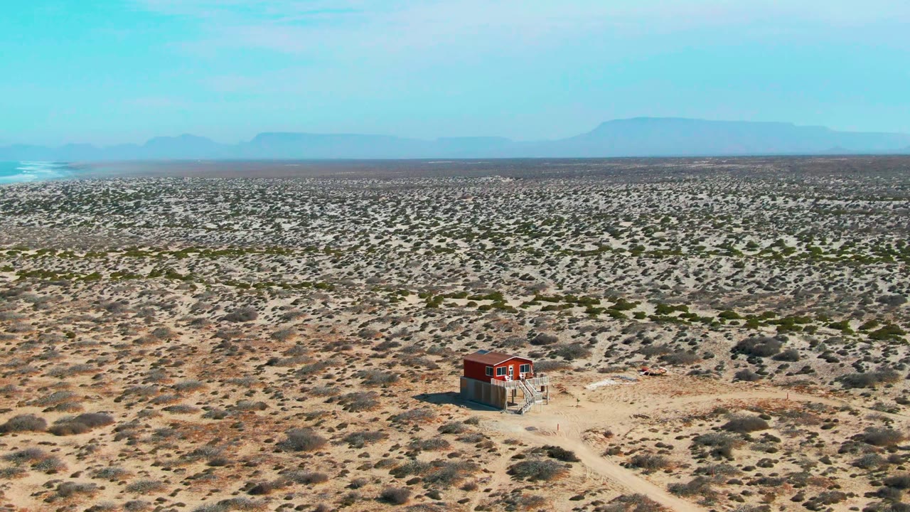 Aerial view over La Bocana desert scrub with a small red structure near the lower center-left, a pale smoke plume at the far left horizon, distant mountains, and no visible people.