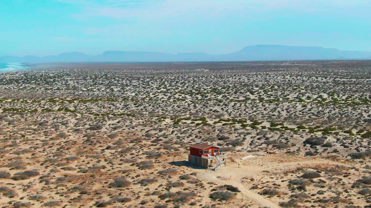 Wide aerial view of scrub desert with a red structure and white smoke near distant mountains.