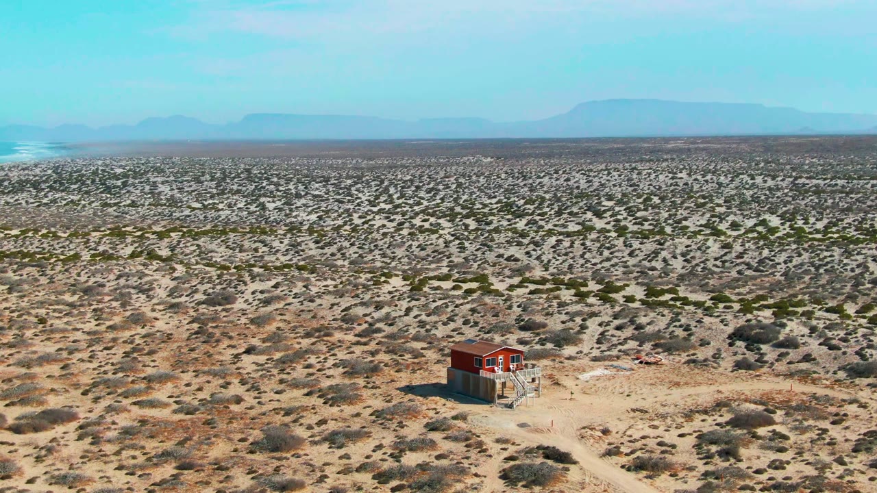 A high view over La Bocana shows a red shack-like structure in the lower right area, pale desert tracks, scrub vegetation, a smoke plume on the left horizon, and no visible people.