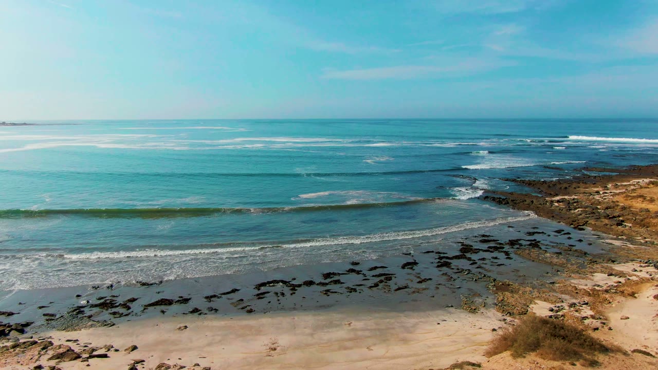 Opening view of La Bocana shoreline with turquoise water, dark rock patches, sandy foreground, dry coastal vegetation, and no visible people.