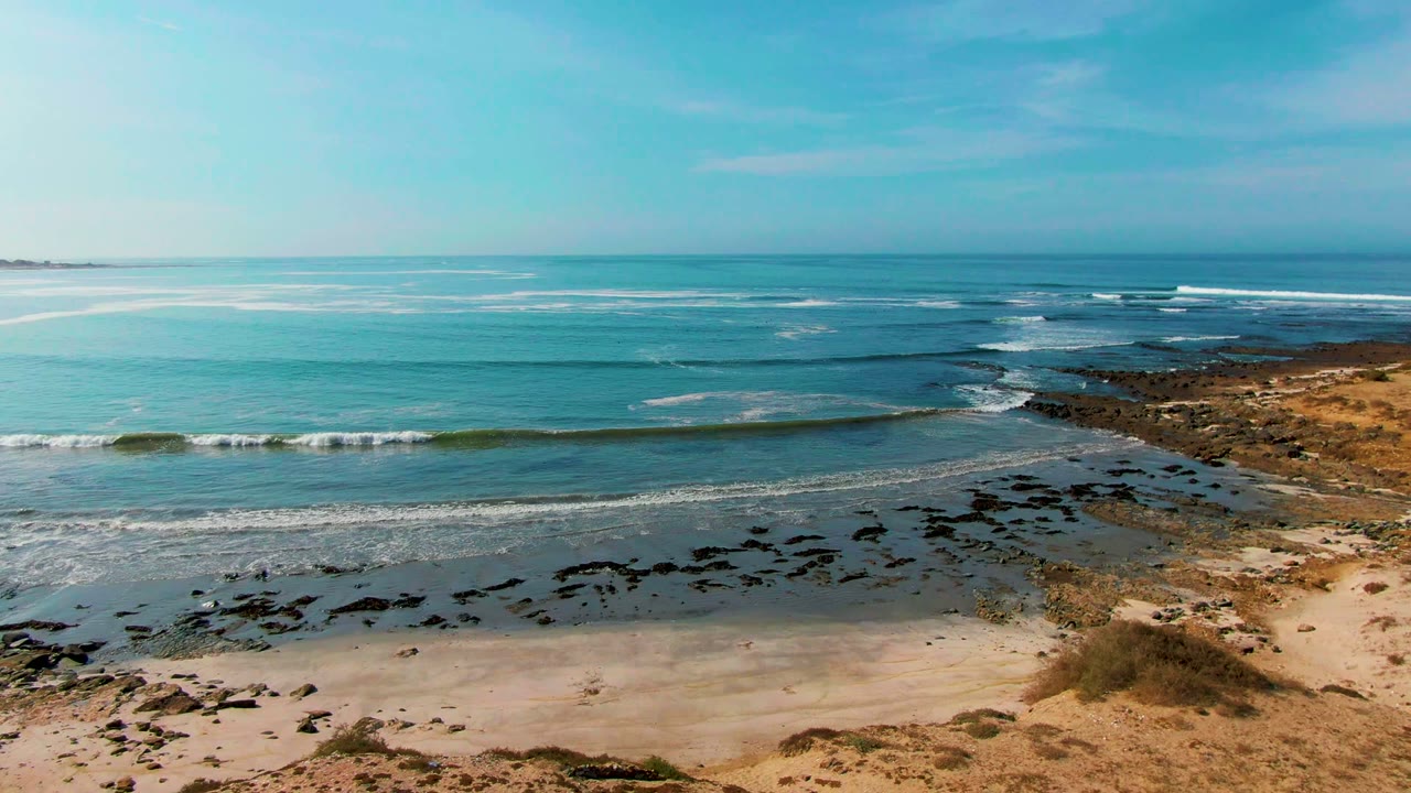 Ocean swell nearing a sandy beach with dark rocks and dry coastal brush.