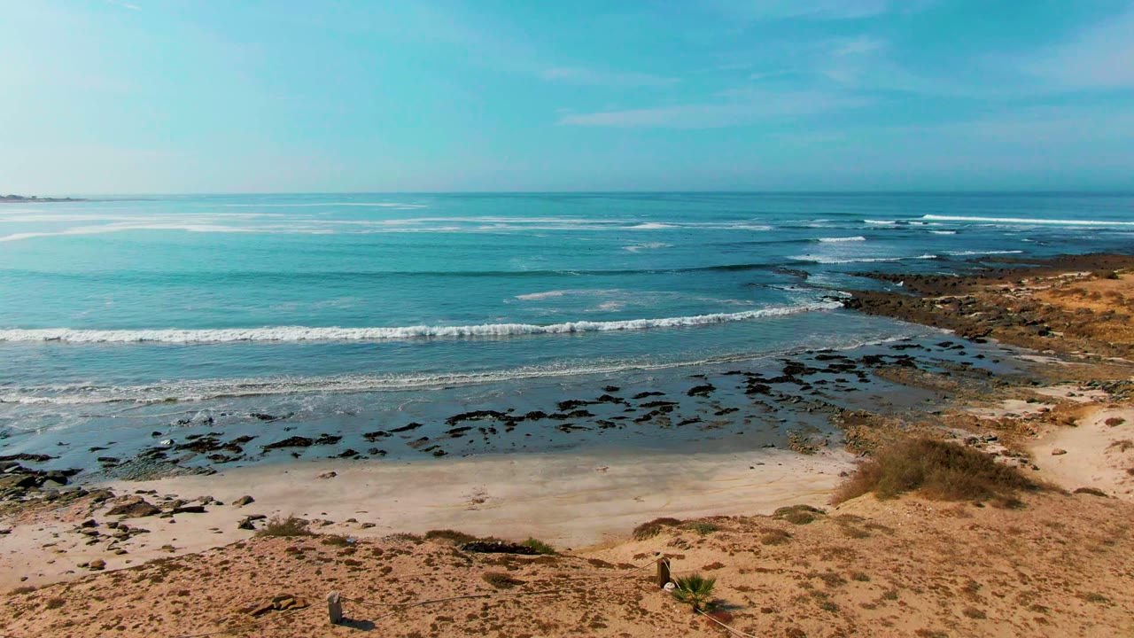 Wide beach and ocean scene with waves, exposed rocks, and coastal scrub.