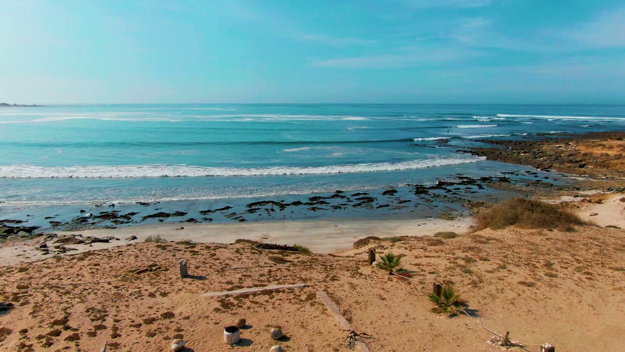 La Bocana beach view with tan sand, scattered rocks, low plants, dark shoreline rocks, blue ocean, clear sky, and no visible people at the opening frame.