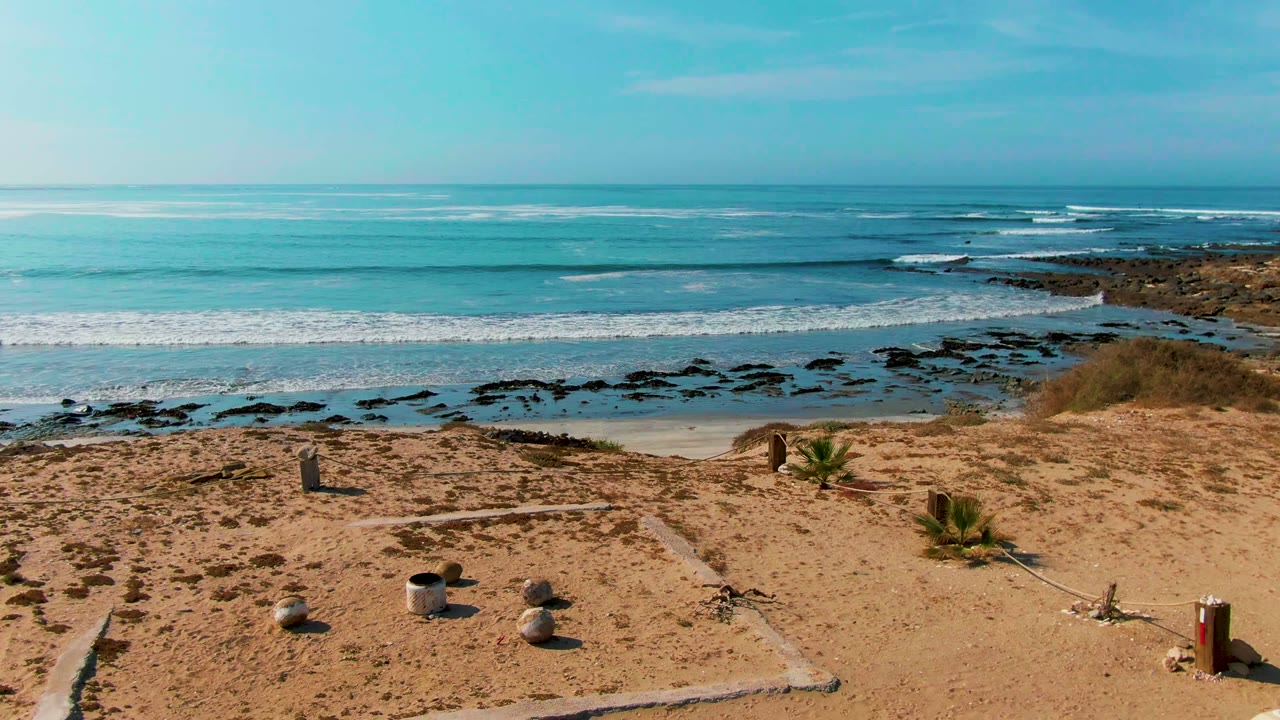 La Bocana coastal view with more open sand in front, a few rocks and plants, white waves breaking across the rocky shallows, and no visible people.