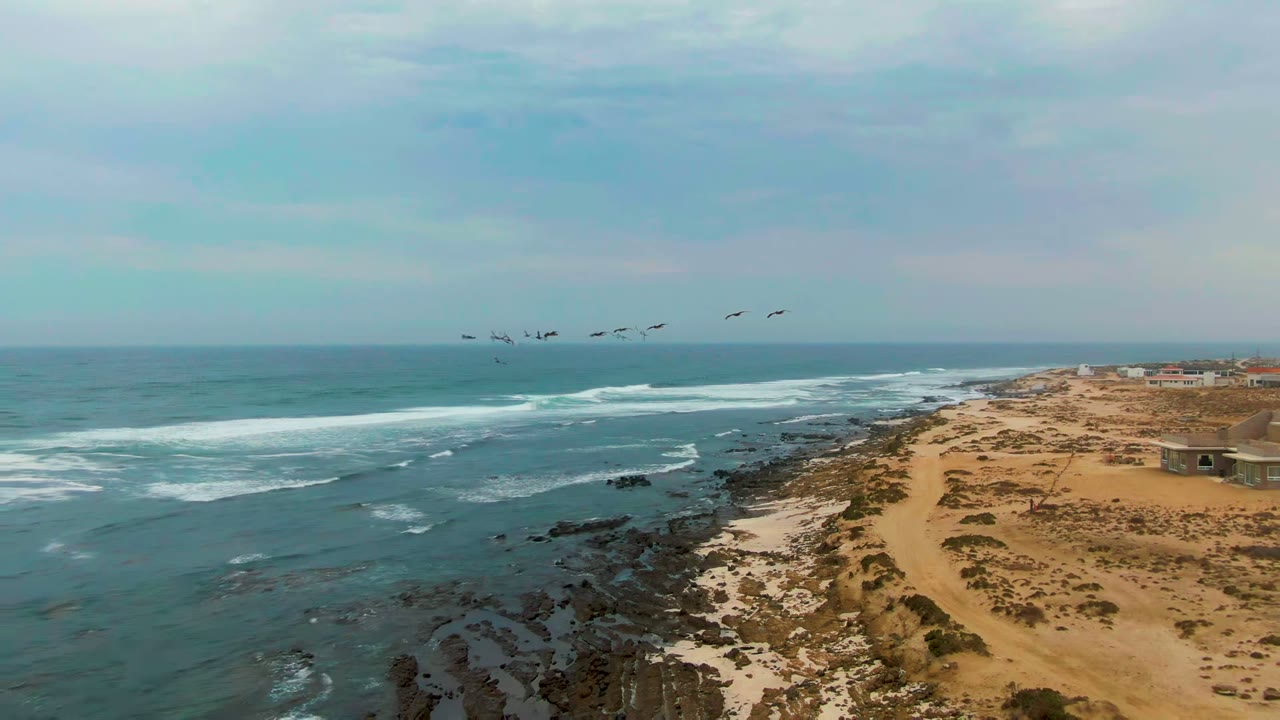 At La Bocana, Baja California Sur, a small flock of birds crosses above teal ocean surf beside a sandy and rocky shoreline, with no visible people.