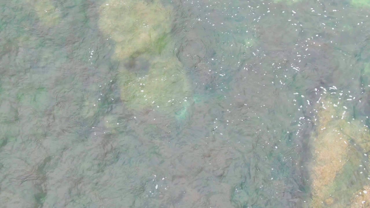 Overhead view of clear shallow water in La Bocana, Baja California Sur, with tan rocks beneath green-blue ripples and no visible people.