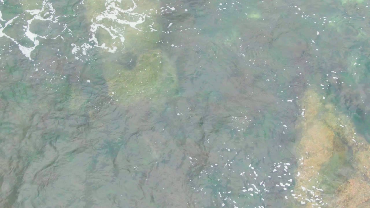 Clear shallow water in La Bocana, Baja California Sur covers a mottled rocky seabed with white foam along the upper left edge and no visible people.