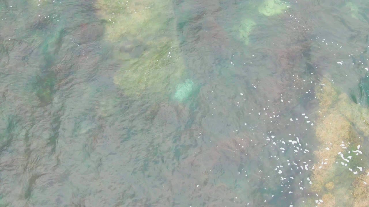 Overhead view of clear blue-green water over submerged rocks in La Bocana, Baja California Sur, with faint foam specks and no visible people.