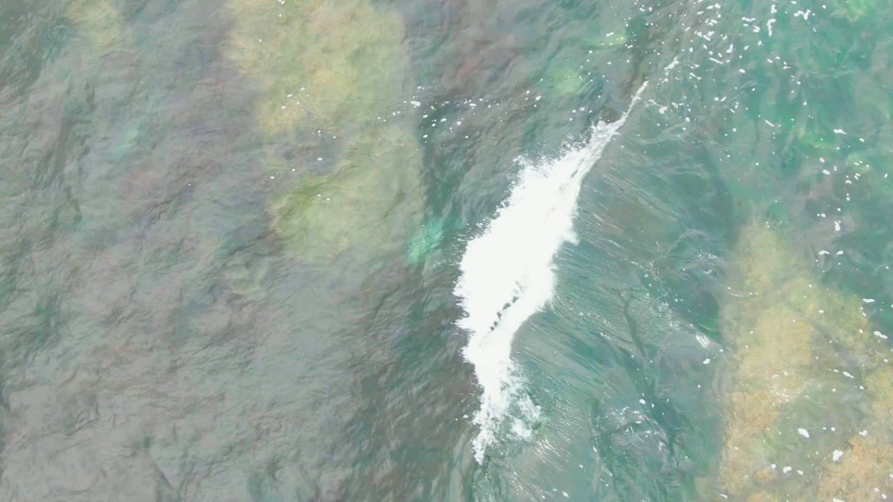 A white foam line enters the overhead La Bocana water frame over blue-green shallows and submerged rock patches, with no visible people.