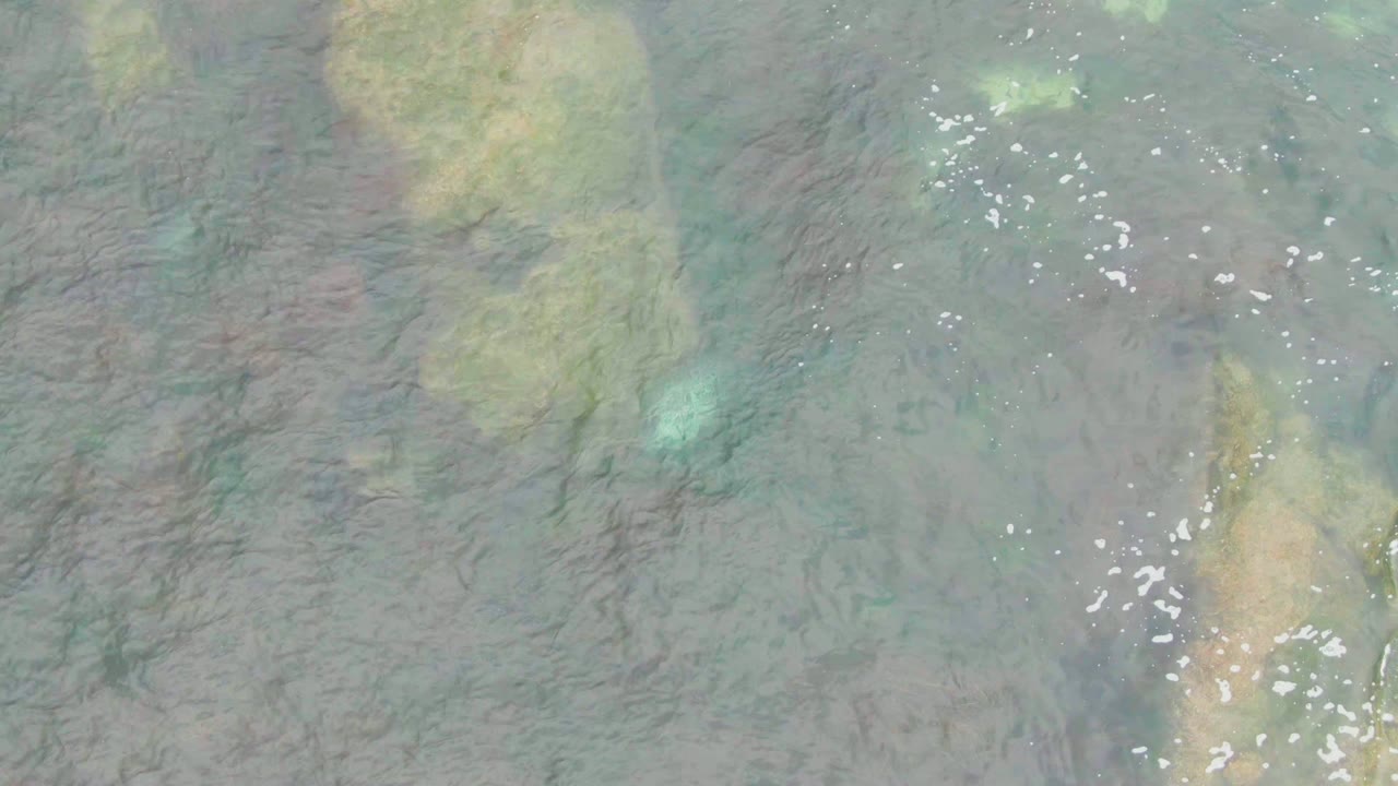 La Bocana coastal water fills the frame with dark rocky lines under the surface, pale green and tan seabed patches, scattered foam flecks, and no visible people.