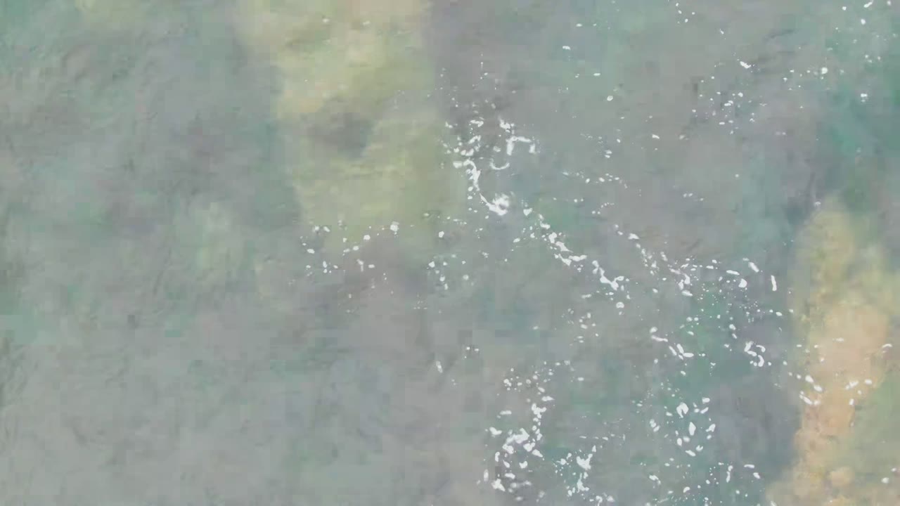 Overhead view of shallow water at La Bocana, Baja California Sur with muted green water, scattered white foam, tan underwater patches, and no visible people.