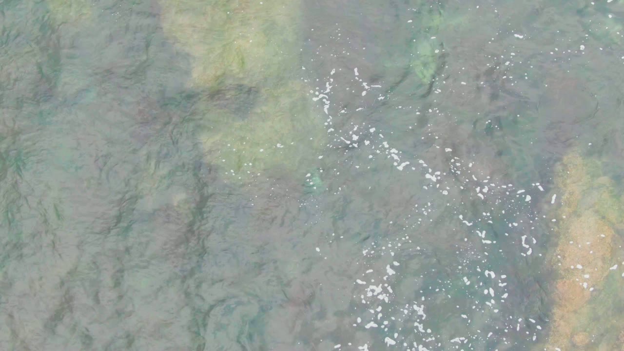 Top-down La Bocana water surface with bright white foam specks, soft blue-green color, darker underwater streaks, and no visible people.