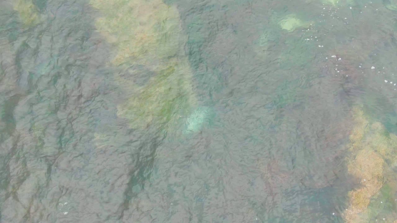 Top-down frame of shallow ocean water in La Bocana, Baja California Sur, Mexico, showing muted green water, brown seabed streaks, pale yellow patches, small foam specks, and no visible people.