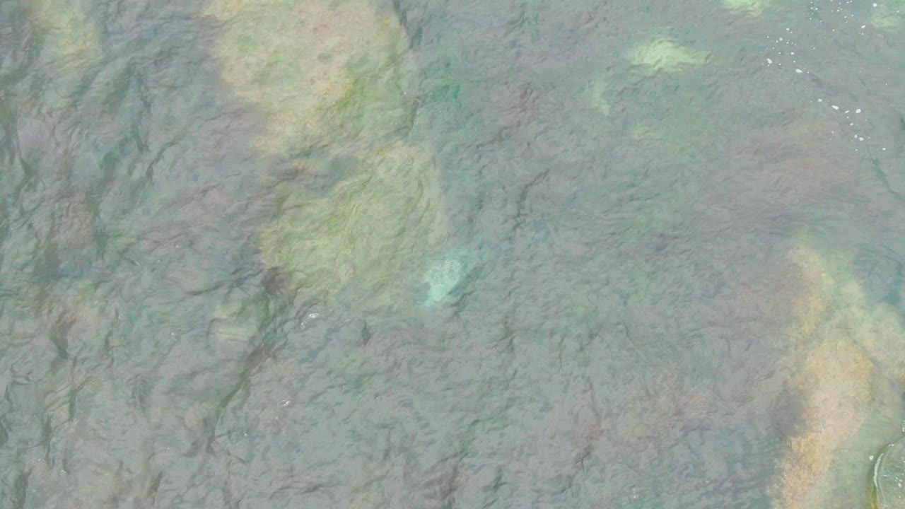 Top-down La Bocana water frame with a turquoise patch near the lower left, yellow-brown seabed areas, dark submerged streaks, light foam flecks, and no visible people.