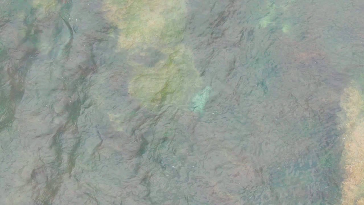 Overhead view of shallow water in La Bocana, Baja California Sur, showing muted blue-green seabed texture with brown rock-like streaks and no visible people.