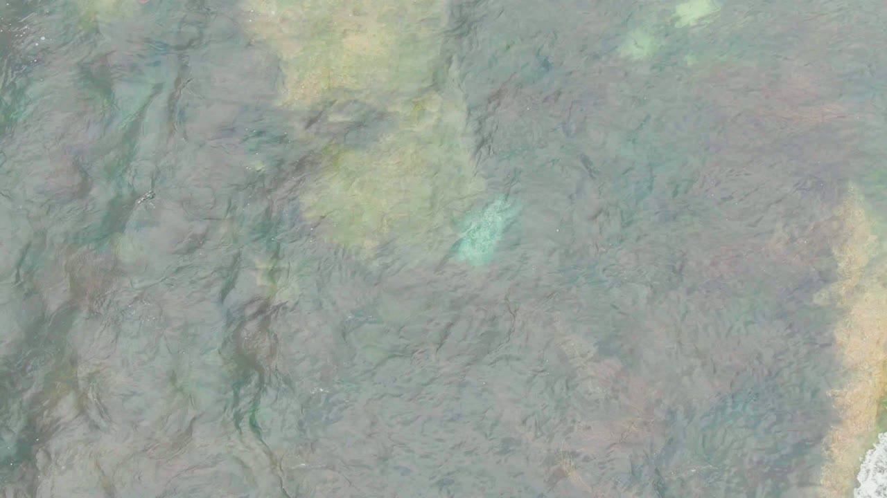 A top-down La Bocana frame shows blue-green shallow water over uneven seabed patterns, with a pale sand-toned band near the right side and no visible people.