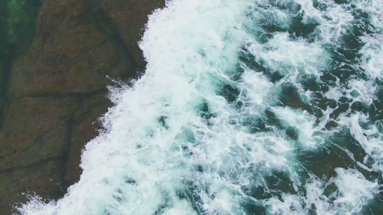 Aerial view in La Bocana, Baja California Sur with white foam pushing over dark and tan rocks while the seal remains visible near the lower left area, with no visible people.