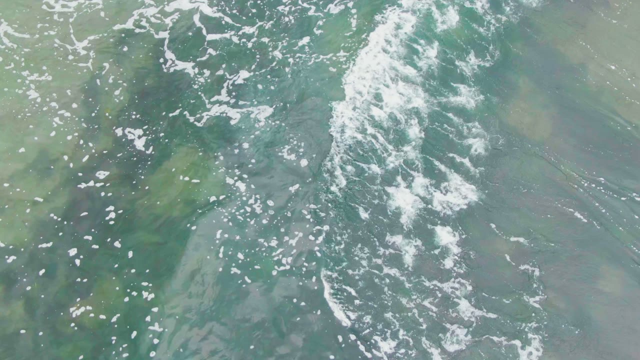 Overhead view of turquoise ocean water in La Bocana, Baja California Sur, with scattered white foam, darker underwater movement near the upper area, and no visible people.
